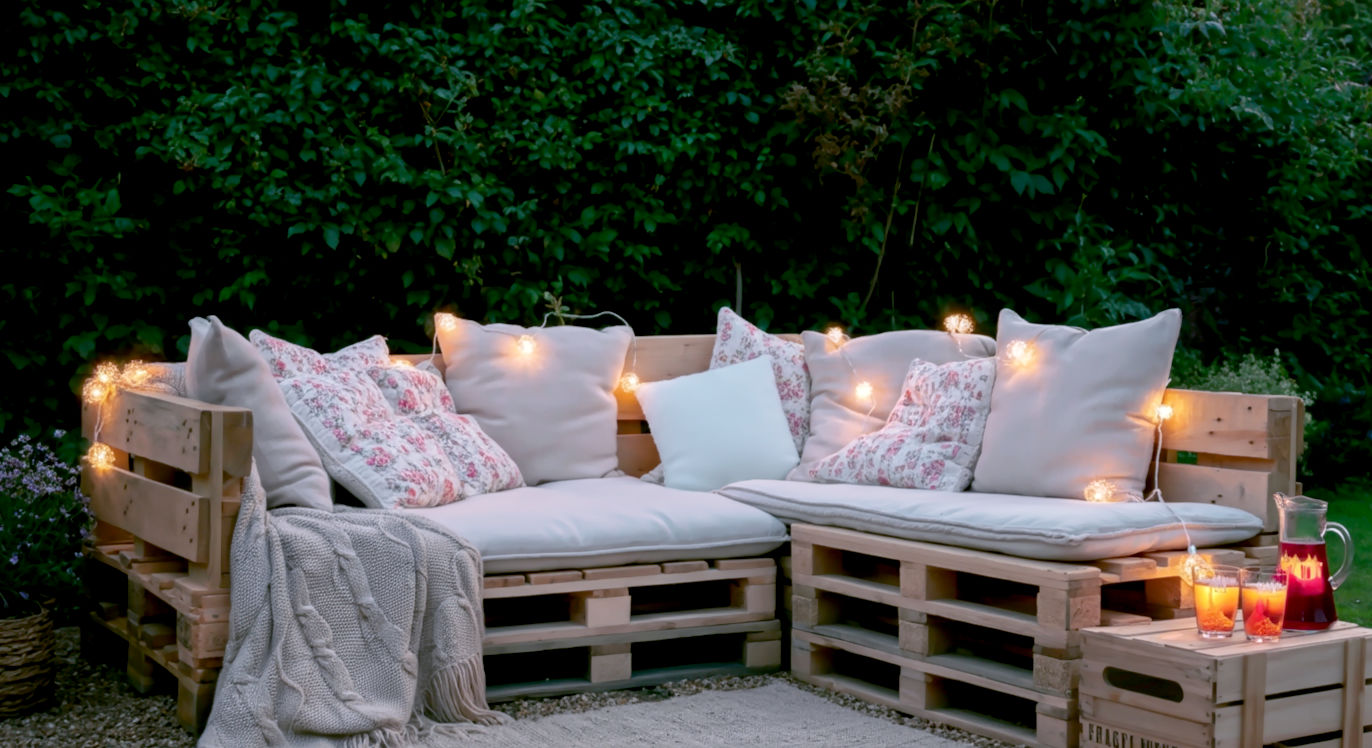 A warm, inviting photograph in the style of Country Living magazine, taken during the 'golden hour'. It features a large, L-shaped pallet wood sofa in a lush, slightly overgrown British garden. The sofa is laden with comfortable, mismatched cushions and a chunky knitted throw. A small, solar-powered fairy light string is woven through the backrest, beginning to glow as dusk approaches. On a small crate table next to it sits a jug of Pimm's and a couple of glasses. The mood is idyllic, relaxed, and quintessentially British summer.