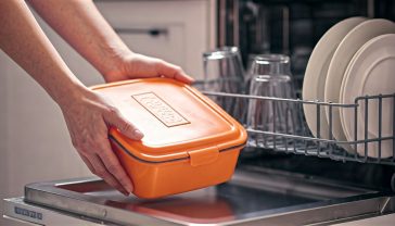 A hyper-realistic, professional photograph in the style of a Good Housekeeping feature. The image shows a pair of hands hesitating to place a brightly coloured plastic lunchbox onto the bottom rack of a modern, clean, stainless-steel dishwasher. The top rack is neatly loaded with sparkling glasses and white ceramic plates. The lighting is bright, clean, and natural, coming from a nearby kitchen window, creating soft shadows. The mood is one of relatable, everyday uncertainty, perfectly capturing the theme of the article. The focus is sharp on the plastic box and the confusing symbols embossed on its base.