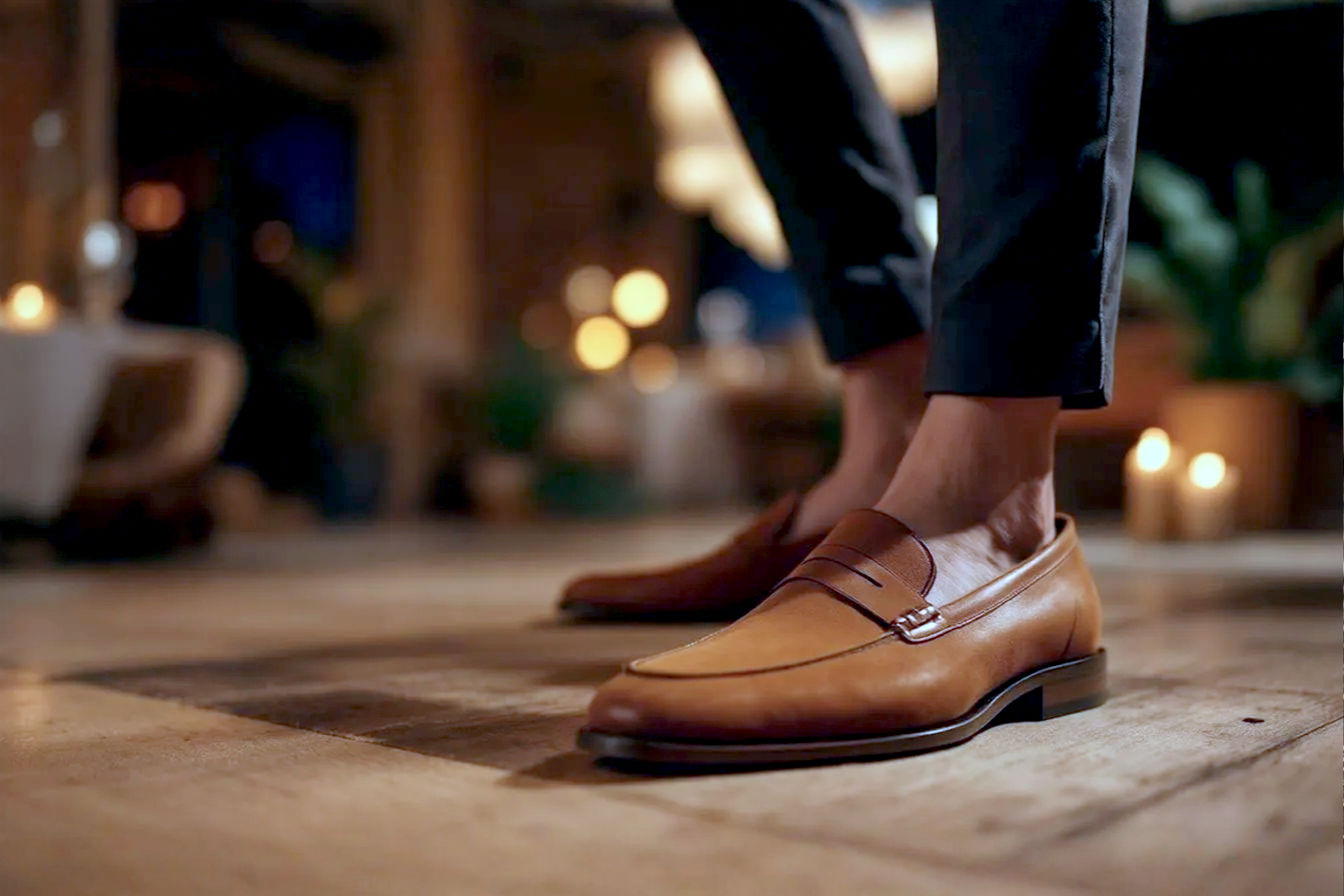 A high-quality, professional photograph featuring a well-dressed person (gender-neutral or showing both men and women) showcasing stylish loafers in a sophisticated semi-formal setting. The person is perhaps standing or taking a step, highlighting the shoes. The background could be a subtly blurred elegant event space, a polished urban street, or a chic interior, conveying a semi-formal ambiance. The loafers should be the focal point, looking clean, polished, and made of luxurious material (leather or suede). Lighting is soft and flattering, emphasizing texture and detail. The overall mood is confident, refined, and approachable.