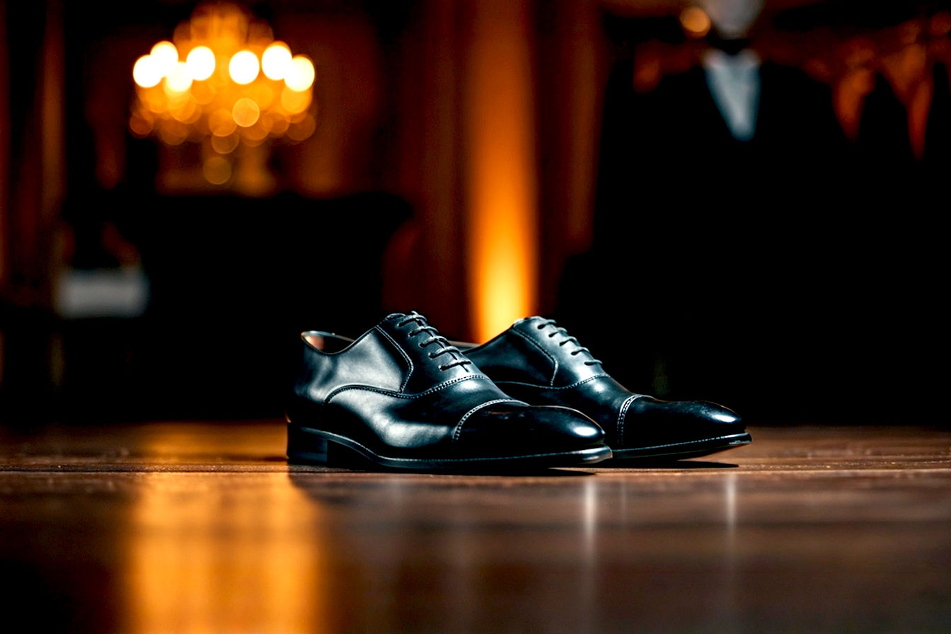 A pair of highly polished, classic black cap-toe Oxford formal shoes resting on a pristine, subtly reflective dark wooden floor, with a soft, elegant out-of-focus background of a formal event (e.g., blurred chandeliers, a glimpse of tailored suit fabric). The lighting is sophisticated and warm, highlighting the gleam of the leather. Professional photography style, lifelike, sharp focus on shoes.