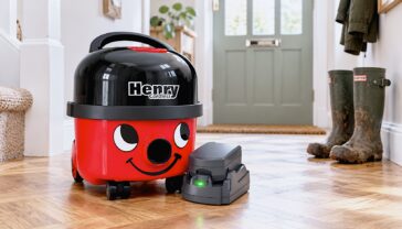 The classic red Henry Cordless vacuum cleaner sitting on a stylish, modern parquet floor in a British hallway. A pair of muddy Wellington boots sit blurred in the background. The green LED light on the battery pack is glowing, indicating it is ready to use.