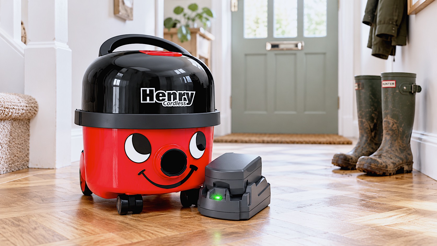 The classic red Henry Cordless vacuum cleaner sitting on a stylish, modern parquet floor in a British hallway. A pair of muddy Wellington boots sit blurred in the background. The green LED light on the battery pack is glowing, indicating it is ready to use.