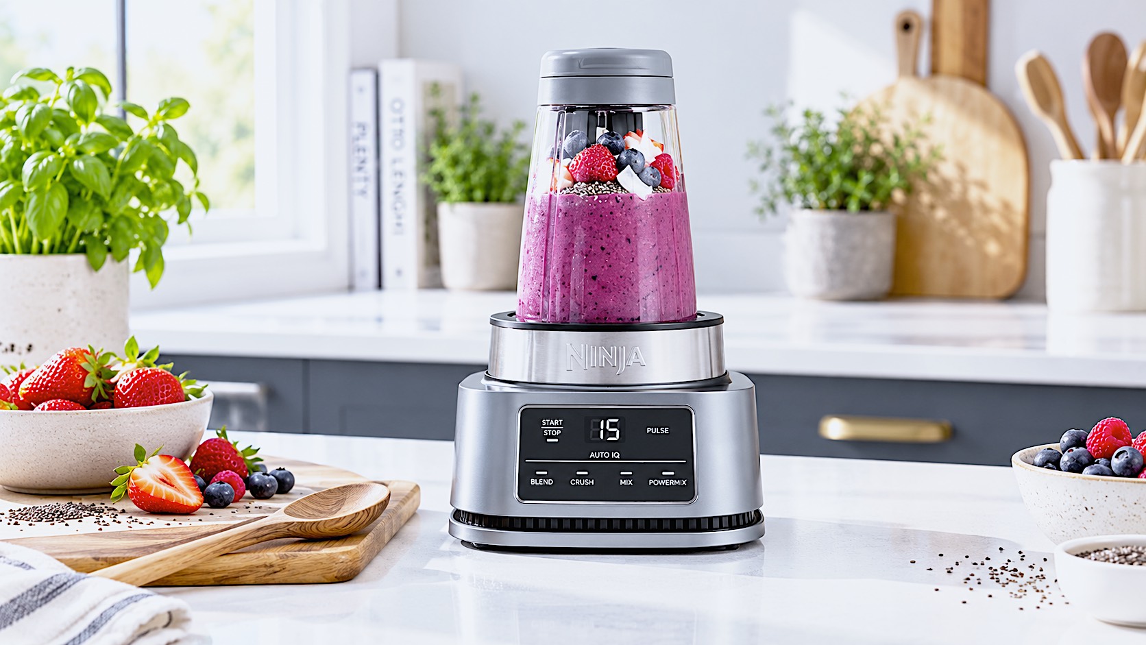 A brightly lit photograph of the silver Ninja Foodi CB100UK blender sitting on a sleek white quartz kitchen worktop in a modern British kitchen. The 400ml Power Nutri Bowl is attached, filled with a vibrant, thick purple açaí and mixed berry smoothie bowl. Fresh strawberries and a wooden spoon rest on a chopping board nearby.