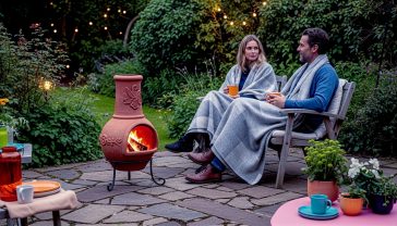 A hyper-realistic, professional photograph of a rustic, terracotta clay chiminea glowing warmly on a flagstone patio in a lush, green British cottage garden during twilight. A cosy couple in their 30s sits nearby on wooden chairs, wrapped in wool blankets, holding mugs of tea. Soft, ambient light from the fire and string lights illuminates the scene. The mood is tranquil, warm, and aspirational. In the style of a high-end feature in Country Living or Gardens Illustrated magazine, with a shallow depth of field focusing on the chiminea.