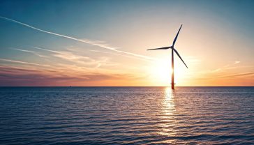 A hyper-realistic, professional photograph in the style of a BBC documentary still. The shot captures a magnificent, modern offshore wind turbine at sunrise, off the coast of Norfolk. The composition is a wide-angle view, showing the turbine's immense scale against the vast sea. The first light of dawn casts a soft, golden glow on the horizon, with calm, deep blue waters below. The sky is filled with a few wispy, pink-tinged clouds. The mood is one of awe, technological prowess, and serene, clean power. The image should feel quintessentially British, evoking a sense of calm optimism and sustainable progress.