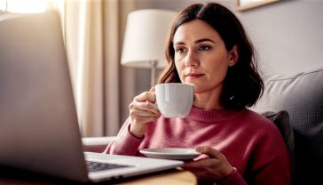 A hyper-realistic, professional photograph showing a thoughtful British person in their late 30s, sitting in a cosy, well-lit living room with a cup of tea. They are looking at a laptop screen with a slightly sceptical but confident expression. The laptop screen is angled towards the viewer but is slightly out of focus, while the person's face is the sharp focus. The mood is one of quiet diligence and empowerment, with soft, natural lighting from a nearby window, evoking a sense of trust and security. Style it like a feature in a high-quality weekend magazine.