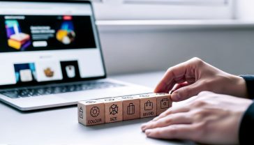 A hyper-realistic, professional photograph in the style of a clean, modern tech blog. The image shows a close-up of a person's hands neatly organising labelled wooden blocks on a minimalist light grey desk. The blocks have simple, clear icons and words like "Colour," "Size," and "Brand." In the background, a sleek laptop screen is slightly out of focus, displaying a vibrant, well-designed WooCommerce store page. The lighting is soft and natural, coming from a window, creating a calm, focused, and organised mood. The overall aesthetic is professional, accessible, and hints at the digital craftsmanship of building a UK-based online shop.