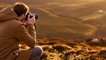 A hyper-realistic, professional photograph. A young person in their late 20s, dressed for a British country walk (e.g., Barbour-style jacket), is kneeling on one knee in the Scottish Highlands during a stunning golden hour. They are holding a DSLR camera, looking intently through the viewfinder, composing a shot of a majestic stag in the middle distance. The scene is bathed in warm, low light, with long shadows stretching across the heather-covered landscape. The mood is one of intense focus, creativity, and connection with nature. Style of a top travel photographer.