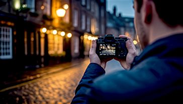 A hyper-realistic, professional photograph capturing the essence of low-light photography in a classic British setting. Subject: A photographer in their late 20s, viewed from behind, holding a modern mirrorless camera up to their eye. They are composing a shot of a rain-slicked, cobbled street in a historic city like York or Edinburgh at dusk. Composition: The photographer is on the right third of the frame, creating a strong focal point. The street stretches away into a soft, blurry background (bokeh) where warm lights from a pub window glow. The reflection of streetlights shimmers on the wet stones in the foreground. Lighting: The scene is lit by the ambient glow of old-fashioned streetlamps and the warm light spilling from the pub, creating a moody, atmospheric feel with deep shadows and soft highlights. The camera's rear screen should be faintly illuminated, showing a hint of the settings. Mood & Style: The mood is contemplative, creative, and slightly nostalgic. The style should be that of a high-end travel magazine feature, with rich colours, sharp focus on the photographer and their camera, and an overall feeling of authenticity. The image should convey the idea that understanding ISO allows you to capture beauty even in challenging British light.