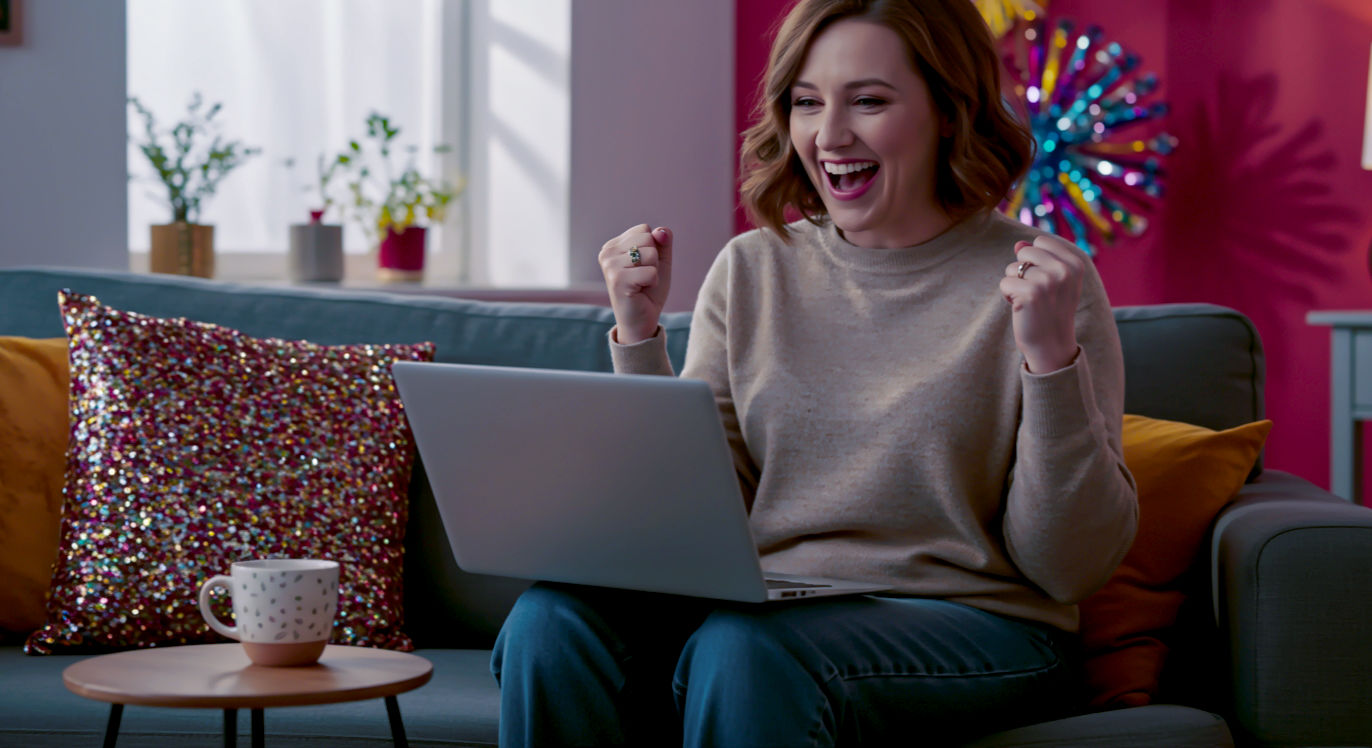 A hyper-realistic, professional photograph in the style of a modern lifestyle magazine. A cheerful British woman in her late 30s is sitting on a comfy sofa in a bright, cosy living room, holding a laptop. She has a huge, joyful smile on her face as she looks at the screen, celebrating a win. A mug of tea sits on a small table beside her. The lighting is soft and natural, coming from a nearby window, creating a warm and aspirational mood. The overall feel is authentic, relatable, and captures the excitement of winning.