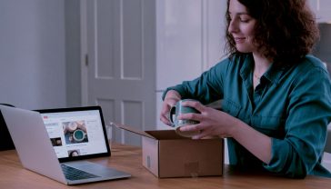 A hyper-realistic, professional photograph in the style of a feature for The Sunday Times. A friendly and determined-looking British woman in her early 30s sits at her kitchen table in a bright, modern London flat. She is smiling slightly as she packs a beautifully designed, handmade ceramic mug into a branded cardboard box, ready for shipping. Her laptop is open next to her, showing an Etsy seller dashboard. The scene is bathed in soft, natural morning light, creating a mood of optimistic, modern entrepreneurship. The composition is a medium shot, with a shallow depth of field focusing on her hands and the product. The overall aesthetic is clean, authentic, and aspirational.