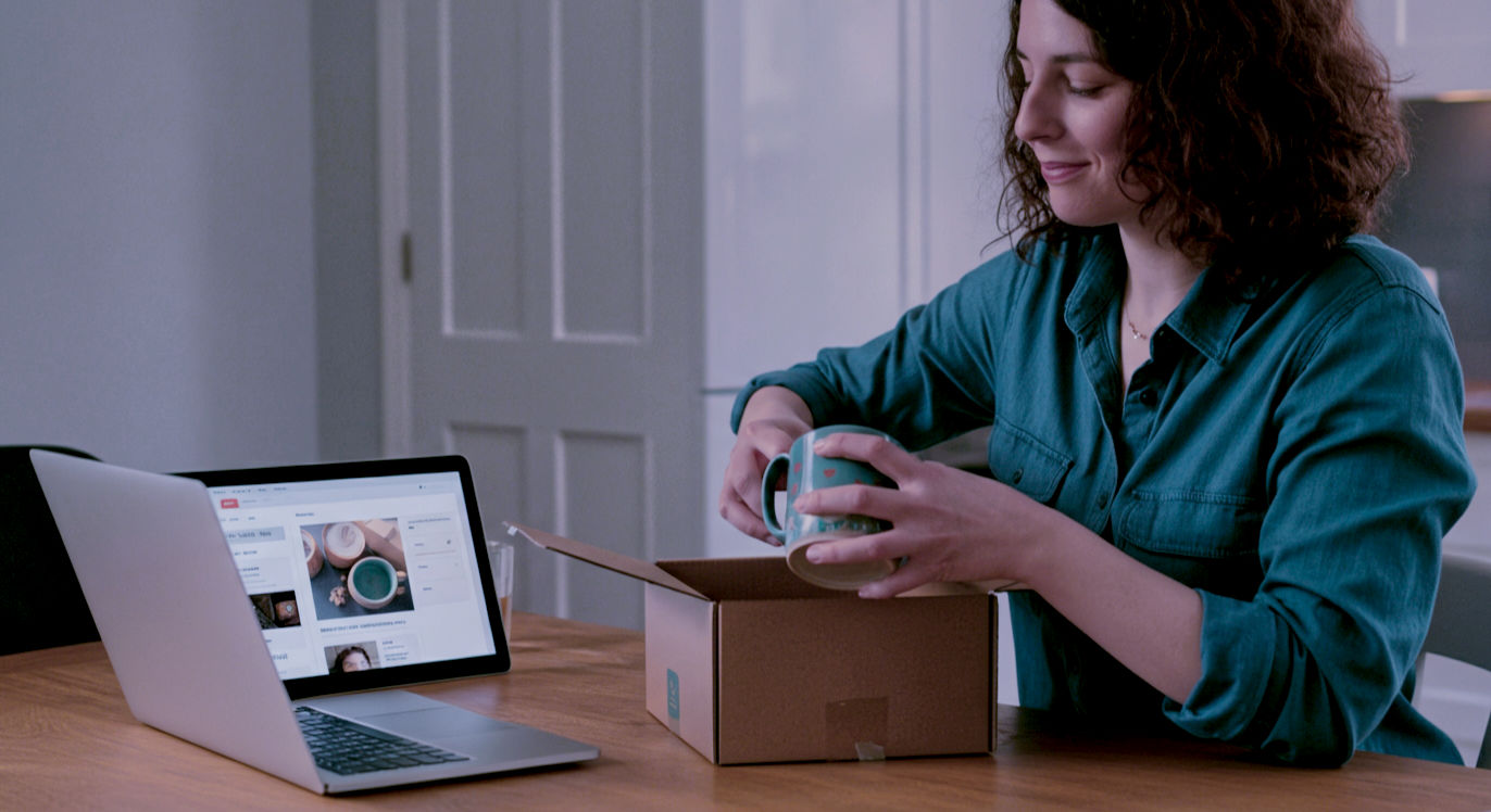 A hyper-realistic, professional photograph in the style of a feature for The Sunday Times. A friendly and determined-looking British woman in her early 30s sits at her kitchen table in a bright, modern London flat. She is smiling slightly as she packs a beautifully designed, handmade ceramic mug into a branded cardboard box, ready for shipping. Her laptop is open next to her, showing an Etsy seller dashboard. The scene is bathed in soft, natural morning light, creating a mood of optimistic, modern entrepreneurship. The composition is a medium shot, with a shallow depth of field focusing on her hands and the product. The overall aesthetic is clean, authentic, and aspirational.