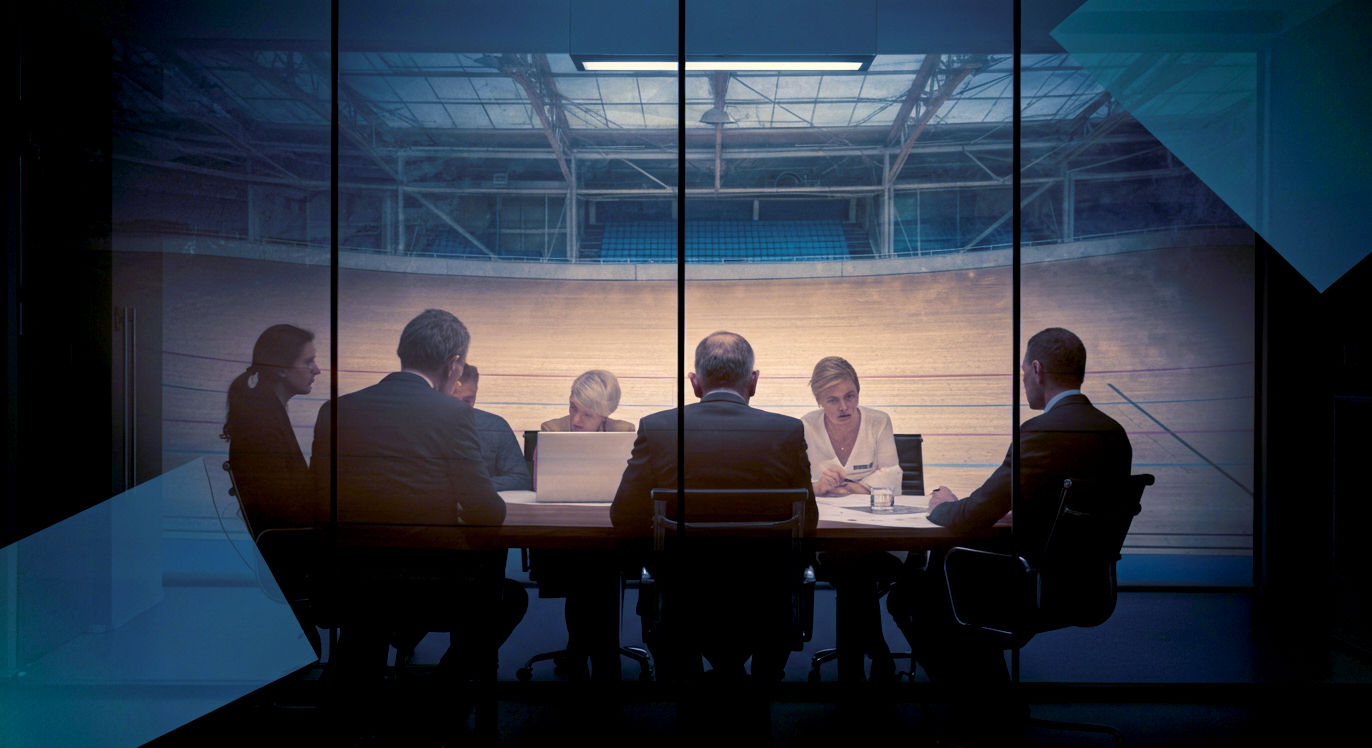 A highly-stylised, professional photograph with a cinematic feel. The composition is a clever double exposure. In the foreground, a group of business executives in a sleek, glass-walled London boardroom are intensely focused on a strategic plan. Reflected in the glass wall behind them is a ghostly, semi-transparent image of the British Cycling team in a velodrome. The lighting is cool and contemporary inside the boardroom, contrasting with a warmer, more dramatic lighting on the historical reflection. The mood is one of legacy, evolution, and the timeless human quest for a competitive edge, connecting Britain's modern business acumen to its sporting heritage.