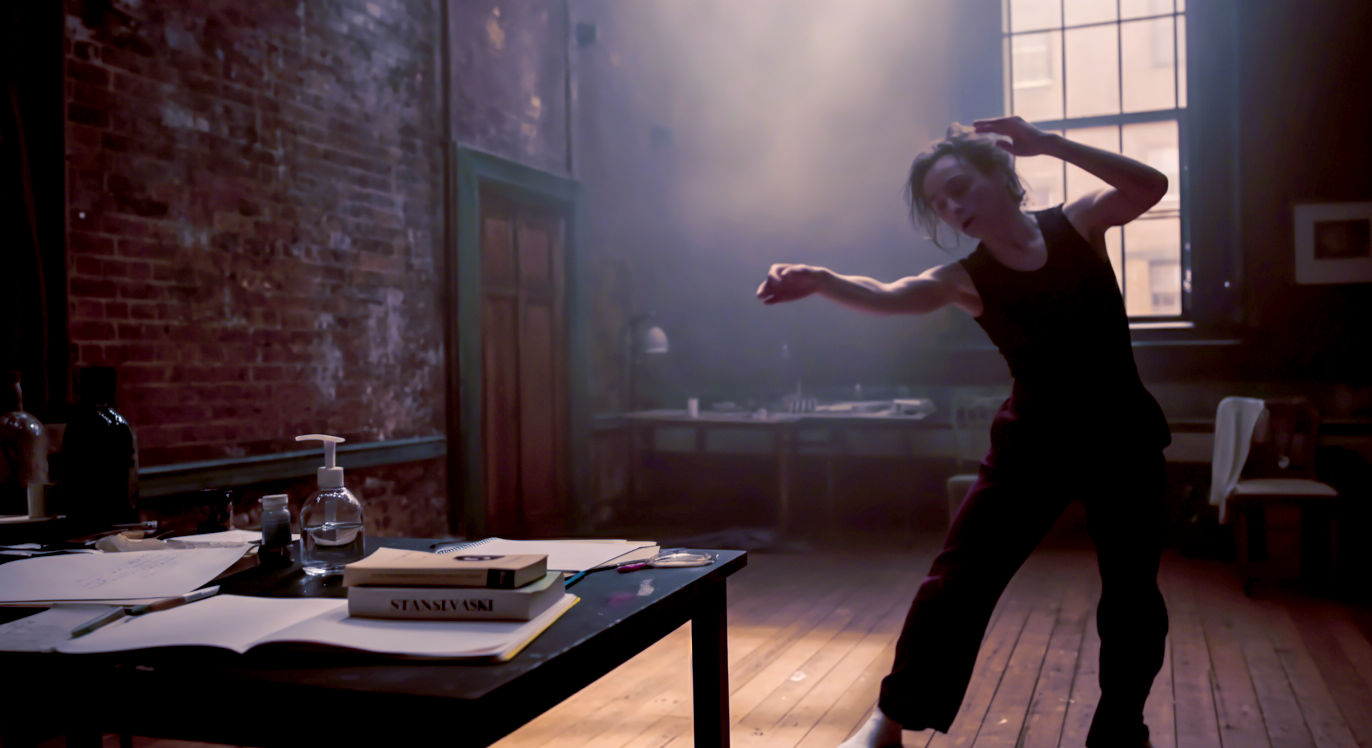 A hyper-realistic, candid photograph in the style of a backstage documentary feature for a publication like The Guardian. The scene is a gritty, well-used London rehearsal room with exposed brick walls and a worn wooden floor. An actor in simple black rehearsal clothes is captured mid-movement, their expression one of intense concentration. In the background, a table is cluttered with a marked-up script, notebooks, and a visible copy of a book by Stanislavski. The lighting is natural, streaming from a large, off-camera window, creating long shadows and highlighting the dusty, authentic atmosphere of creative work.