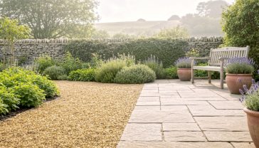 A hyper-realistic photograph in the style of a Gardens Illustrated magazine feature. A beautiful, established British garden in early summer. A winding gravel path made of golden Cotswold chippings leads past a lush herbaceous border towards a stone patio. On the patio, a comfortable wooden bench sits next to a large terracotta pot planted with lavender. In the background, a low dry stone wall, covered in creeping thyme, separates the garden from a gentle green hill. The lighting is soft and warm, like a hazy afternoon sun, creating a peaceful and aspirational mood. The focus is sharp on the texture of the stone path and wall.