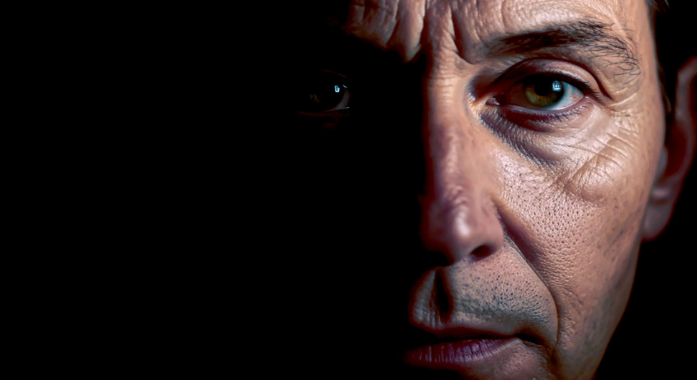 A hyper-realistic, intimate close-up photograph, reminiscent of a cinematic character study. The subject is an actor's face, mid-40s, with expressive lines. Half of the face is softly lit by a single key light, revealing a complex emotion of quiet realisation in one eye, while the other half is in deep, contemplative shadow. The composition is tightly cropped and slightly off-centre, with a sharp focus on the illuminated eye. The mood is raw, profound, and deeply human, captured in a painterly palette of warm skin tones against a near-black background.