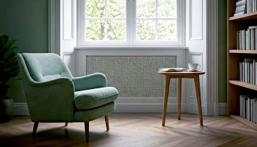 A hyper-realistic photograph capturing the essence of a serene and mindful British living room. The shot should be from a low angle, looking across a cosy, uncluttered space bathed in soft, natural light streaming through a large sash window. To the left, a comfortable, well-loved armchair in a muted sage green sits next to a small wooden side table holding a single cup of tea and an open book. In the background, there are shelves with a curated mix of books and healthy green houseplants. The overall mood is calm, peaceful, and restorative, with a colour palette of soft greens, warm woods, and gentle creams, evoking a modern yet timeless British sanctuary. Style it like a feature in The Sunday Times Style magazine.
