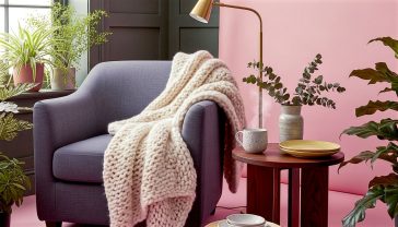 Professional interior photography, in the style of Elle Decoration UK. The subject is a beautifully styled corner of a contemporary British living room. The main focus is a comfy, deep-grey linen armchair draped with a chunky, cream-coloured wool throw. Next to it, a dark wooden side table holds a textured ceramic mug steaming gently and a small vase with dried eucalyptus branches. In the background, there's a hint of a dark green, subtly panelled wall and a sleek brass reading lamp. The lighting is soft, natural light coming from a large window just out of frame, creating gentle shadows that highlight the different textures of the linen, wool, wood, and ceramic. The mood is cosy, sophisticated, calm, and inviting.