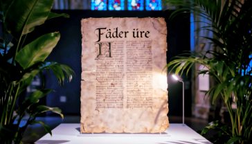 A hyper-realistic photograph in the style of a museum exhibit. An ancient, weathered manuscript page featuring the Old English Lord's Prayer ('Fæder ūre') is illuminated by a soft, single light source from the side. In the soft-focus background, a stained-glass window from a classic English church is visible, casting faint, coloured light. The mood is reverent, historical, and scholarly, with a rich, dark palette emphasizing the texture of the vellum and the faded ink.