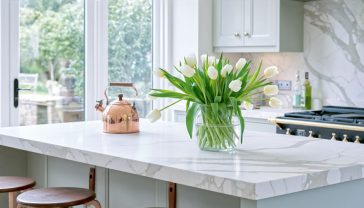 Professional, magazine-style photograph of a bright, modern British kitchen with a large central island topped with a stunning slab of Calacatta Gold marble. A copper kettle sits on the worktop next to a vase of fresh white tulips. The lighting is soft and natural, coming from a large window overlooking a green garden, creating a feeling of calm, aspirational luxury. The style should be reminiscent of a feature in 'Livingetc' or 'House & Garden'.