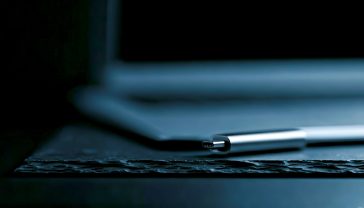 A hyper-realistic, professional product photograph styled for a modern tech blog. A sleek, metallic dual-connector USB-C and USB-A flash drive rests on a minimalist, textured slate surface. One end is plugged into the USB-C port of a modern, ultra-slim laptop (like a MacBook Air or Dell XPS), which is slightly out of focus in the background. The scene is lit with soft, cool, directional studio lighting that highlights the brushed metal texture of the drive and creates subtle, elegant reflections. The overall mood is sophisticated, clean, and professional, conveying speed and reliability.