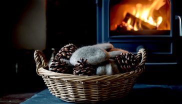 A hyper-realistic, professional photograph in the style of a feature in Country Life magazine. The shot is a close-up, focusing on a hand-woven wicker basket filled with natural wood wool firelighters and wax-dipped pinecones. The basket sits on a rustic slate hearth next to a cast-iron wood-burning stove, in which a gentle flame is just beginning to catch. The lighting is warm and golden, as if from the fire itself, creating a cosy, inviting, and quintessentially British atmosphere. The mood is one of sustainable comfort and quiet tradition.
