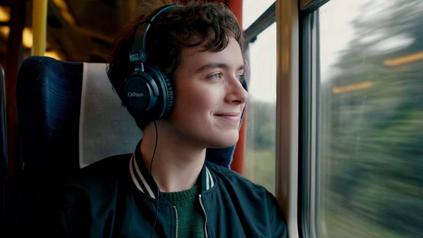 A hyper-realistic, professional photograph capturing the joy of offline music in a classic British setting. A young person in their early 20s, wearing stylish headphones (like Bose or Sony), is sitting on a Chiltern Railways train, looking out of the window at the beautiful, green English countryside rolling by. They have a subtle, happy expression. The lighting is soft and natural, streaming through the train window, creating a warm and slightly nostalgic mood. The composition is a medium shot, focusing on the person but keeping the quintessentially British train interior and scenery clearly visible. The overall aesthetic is clean, modern, and relatable, in the style of a high-end lifestyle magazine.