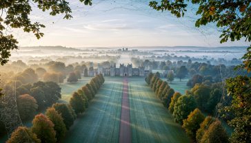A hyper-realistic, professional photograph in the style of a feature for Country Life magazine. The view is an aerial shot on a soft, slightly misty autumn morning, looking across Windsor Great Park. The composition balances grand architecture with the natural, rolling landscape of British parkland. The lighting is the gentle, diffused light of early morning, creating a serene and timeless mood that evokes heritage, privacy, and regal splendour.