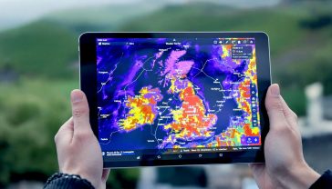 A high-angle, lifelike professional photograph of an iPad displaying a vibrant, animated weather radar map. The iPad is held by a person with a subtle background suggesting a UK outdoor setting (e.g., a misty coastline, a rolling green hill, or a city skyline). The image should convey a sense of preparedness and modern technology, with soft, natural lighting and a slightly shallow depth of field to keep the iPad and its screen as the focal point.