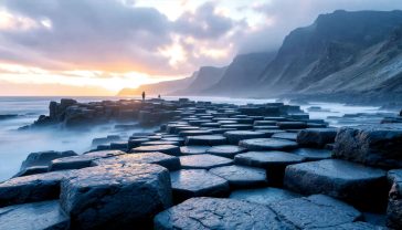 A hyper-realistic, professional photograph in the style of a National Geographic feature. The image captures the Giant's Causeway at sunrise, with the hexagonal basalt columns leading from the foreground into a misty Atlantic Ocean. The first light of dawn casts a warm, golden glow on the wet, dark grey stones, highlighting their geometric patterns. In the background, the dramatic cliffs of the Antrim coast are partially shrouded in a soft sea mist. The composition is wide and epic, emphasising the scale and raw, natural beauty of the landscape. The mood is one of awe, magic, and ancient wonder.