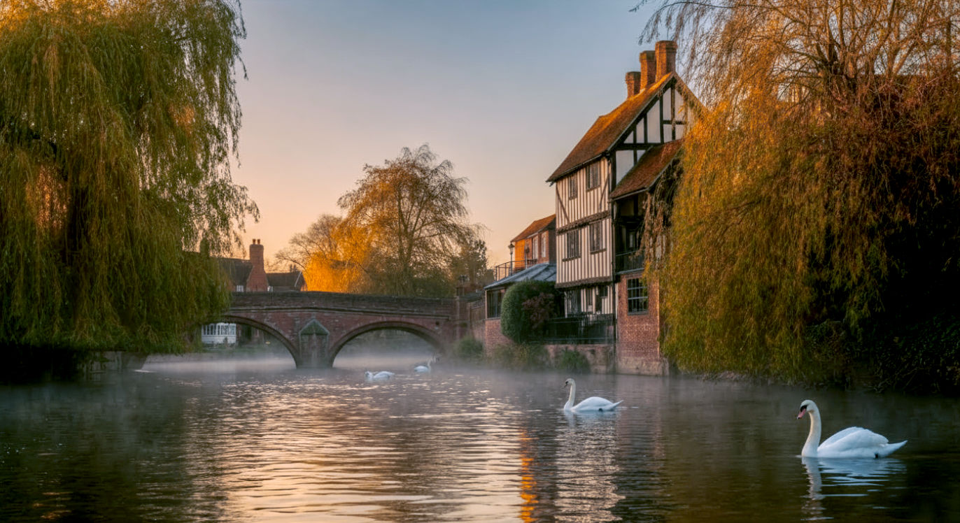A professional, high-resolution photograph of the River Avon in Stratford-upon-Avon at dawn. The Royal Shakespeare Theatre is visible on the riverbank, with its modern tower contrasting with the Tudor-style buildings nearby. A couple of swans glide on the misty water, and the soft morning light gives everything a golden, magical glow. The style should be that of a National Geographic travel photo, capturing both heritage and natural beauty with exceptional clarity and composition.