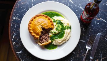 A hyper-realistic, professional food photograph capturing the essence of a traditional London pie and mash meal. The shot is a top-down perspective of a classic white ceramic plate centred on a dark, worn marble tabletop. On the plate sits a golden-brown, flaky pastry pie, slightly broken open to reveal a rich minced beef filling. Next to it is a perfectly smooth scoop of white mashed potato, and generously ladled over everything is a vibrant, bright green parsley liquor. A bottle of chilli-infused malt vinegar sits just in the frame. The lighting is soft and natural, reminiscent of daylight coming through a traditional shop window, creating gentle shadows that highlight the textures of the food. The mood is warm, comforting, and deeply nostalgic, evoking a sense of authentic British culinary heritage.