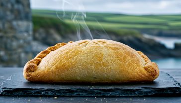 A hyper-realistic, professional food photograph in the style of a BBC Good Food magazine feature. The image shows a perfectly baked, golden-brown Cornish pasty resting on a piece of rustic slate. The pasty is D-shaped with a thick, beautifully crimped edge on the side. A small wisp of steam is gently rising from a small slit in the crust. In the soft-focus background, hint at a rugged Cornish coastal scene with grey stone walls and a touch of green landscape under a slightly overcast sky. The lighting should be soft and natural, coming from the side to highlight the texture of the pastry. The mood is warm, authentic, and evocative of British heritage.