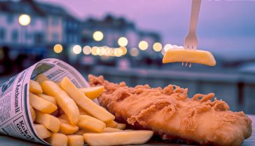 A hyper-realistic, professional photograph capturing the quintessential British fish and chips experience. Subject: A generous portion of golden, crispy battered cod sits alongside a pile of thick-cut, fluffy chips, served in an open newspaper-print cone. A wooden chip fork is stuck into a chip. In the soft-focus background, the warm, inviting lights of a traditional British seaside town at dusk are visible. Style: Warm, nostalgic, slightly rustic, with shallow depth of field. Lighting: Soft, golden-hour light that highlights the texture of the batter and chips. Mood: Comforting, traditional, and quintessentially British.