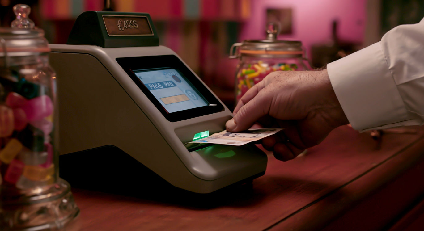 A hyper-realistic, professional photograph showing the hands of a British shopkeeper, slightly weathered, inserting a new £20 polymer banknote into a sleek, modern banknote verifier on a wooden shop counter. The machine's screen glows green with a 'PASS' symbol. In the soft-focus background, we see a classic British retail environment—perhaps a bell jar with sweets or a traditional till. The lighting is warm and inviting, conveying a sense of security, professionalism, and trust.