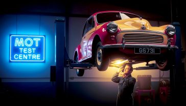 A hyper-realistic, professional photograph capturing a classic 1960s British car, like a Morris Minor, on a hydraulic lift inside a vintage-style garage. A mechanic in classic overalls is thoughtfully inspecting the undercarriage. The iconic blue and white "MOT Test Centre" sign is visible in the background. The lighting is slightly dramatic, with soft, warm light illuminating the car, evoking a sense of nostalgia and British motoring heritage. The style should be reminiscent of a high-quality historical documentary photo.