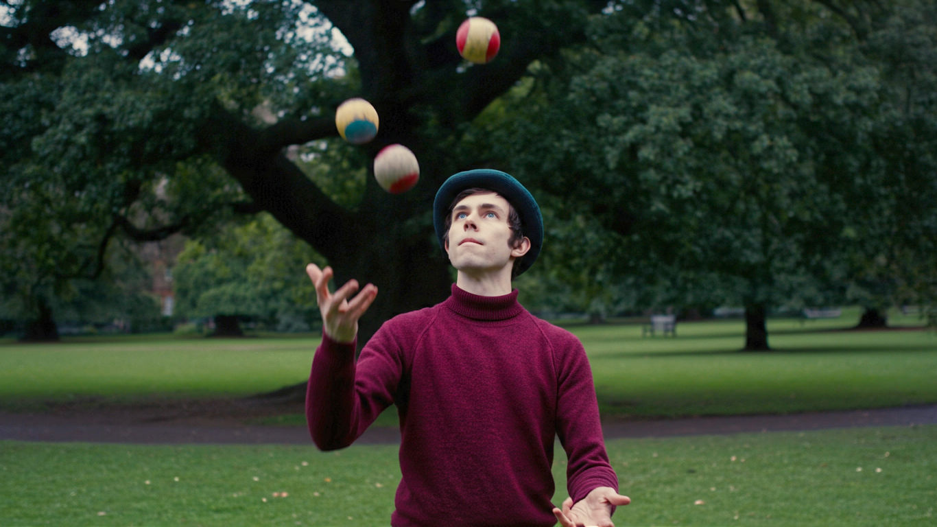 A hyper-realistic, professional photograph in the style of a candid Guardian feature. The subject is a young person in their early twenties, standing in a classic British park like Regent's Park or a Glasgow green. They are mid-juggle with three worn, colourful 'thud' juggling balls, caught in a moment of deep, relaxed concentration. The lighting is soft, overcast British daylight, creating gentle shadows. The background is slightly out of focus, showing green grass, mature oak trees, and perhaps a Victorian-era park bench. The mood is calm, focused, and joyful, capturing the simple, meditative pleasure of the hobby rather than a flashy performance. The composition is a medium shot, framing the juggler from the waist up to capture the full pattern of the balls in the air.