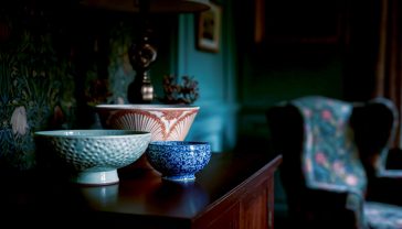 A hyper-realistic photograph in the style of a World of Interiors magazine shoot. A curated collection of decorative bowls is artfully arranged on a dark oak sideboard in a British home. In the centre is a large, vibrant Clarice Cliff Art Deco bowl. To its left, a minimalist, textured stoneware bowl by a contemporary British potter. To its right, a delicate, blue and white Wedgwood porcelain bowl. Soft, natural light from a nearby window illuminates the textures and colours. The background is slightly out of focus, showing a glimpse of a William Morris wallpaper and a cosy armchair. The mood is sophisticated, timeless, and full of character.