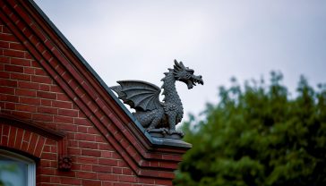 A hyper-realistic, professional photograph in the style of a feature for The World of Interiors magazine. The image captures the detailed craftsmanship of a weathered, dark grey terracotta dragon finial perched on the gable ridge of a Victorian red-brick house in a leafy London suburb. The lighting is soft, overcast daylight, typical of a British afternoon, which highlights the intricate scales and textures of the dragon. The composition is a tight, elegant shot from a slightly low angle, looking up at the finial against a background of soft-focus green trees and a classic lead-grey sky. The mood is one of quiet heritage, enduring craftsmanship, and quintessentially British architectural character.