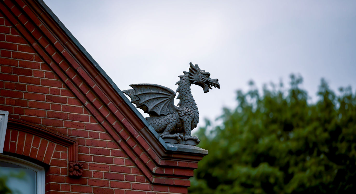 A hyper-realistic, professional photograph in the style of a feature for The World of Interiors magazine. The image captures the detailed craftsmanship of a weathered, dark grey terracotta dragon finial perched on the gable ridge of a Victorian red-brick house in a leafy London suburb. The lighting is soft, overcast daylight, typical of a British afternoon, which highlights the intricate scales and textures of the dragon. The composition is a tight, elegant shot from a slightly low angle, looking up at the finial against a background of soft-focus green trees and a classic lead-grey sky. The mood is one of quiet heritage, enduring craftsmanship, and quintessentially British architectural character.