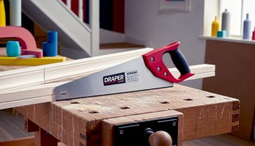 A hyper-realistic, professional photograph in the style of a workshop feature for a British magazine like The English Home. The subject is the Draper Venom handsaw resting on a rustic, solid oak workbench. The workbench is slightly dusty with fine sawdust. In the background, a piece of primed pine skirting board with a freshly cut, perfectly clean 45-degree mitre angle is visible. The lighting is soft and natural, coming from a nearby window, highlighting the saw's red handle and the sharp, gleaming teeth of the blade. The mood is authentic, practical, and aspirational, evoking a sense of British craftsmanship.