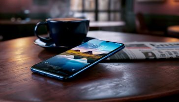 A hyper-realistic, professional product photograph showing the Samsung Galaxy A15 in the "Blue Black" colour. The phone is angled slightly to showcase its flat edges and the "Key Island" feature. It rests on a rustic wooden table in a cosy British cafe, next to a steaming cup of tea and a copy of The Guardian newspaper slightly out of focus in the background. The phone's brilliant Super AMOLED screen is on, displaying a vibrant picture of the Scottish Highlands, highlighting its colour and clarity. The lighting is soft and natural, coming from a nearby window, creating a warm, inviting, and authentically British atmosphere. The style should be clean, high-end, and relatable, like a photo from a TechRadar or Wired UK review.