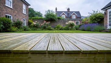 A hyper-realistic, professional photograph in the style of a garden design magazine. The image shows a newly built, handsome wooden deck made from tanalised timber in a classic British garden setting. The timber has a subtle greenish-brown hue, and rain beads on its surface, highlighting its weather-resistant quality. In the background, a lush green lawn, colourful flowerbeds with roses and lavender, and a traditional brick house under a soft, overcast sky create a quintessential UK scene. The composition is a low-angle shot, making the deck look sturdy and inviting, evoking a sense of durability, quality craftsmanship, and lasting value.
