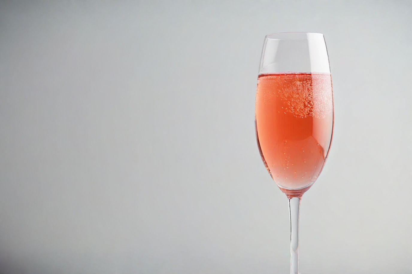 Professional photography of sparkling rosé wine, close-up of a crystal tulip glass with delicate pink bubbles rising through the liquid, soft natural lighting, shallow depth of field, clean white background, droplets of condensation on glass, elegant composition, wine photography style, product photography, high-resolution, advertising quality, 50mm lens, f/2.8, soft shadows