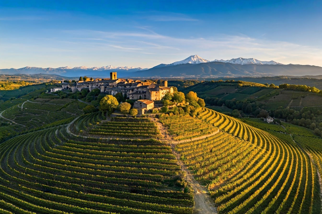 A sweeping aerial photograph capturing the dramatic landscape of the Sud-Ouest wine region during golden hour. In the foreground, terraced vineyards cascade down limestone slopes, with ancient stone farmhouses nestled among the vines. The middle ground reveals a medieval village perched on a hilltop, while the background showcases the majestic Pyrenees mountains with snow-capped peaks catching the sunset light. The image conveys both the rugged beauty and the human heritage of this historic wine region.
