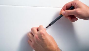 A close-up, lifelike professional photograph of a hand meticulously applying a repair compound to a hairline crack on a glossy white ceramic tile. The background is softly blurred, showing hints of a modern, clean bathroom or kitchen environment.