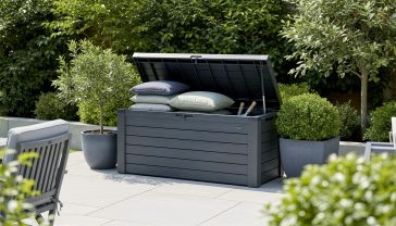 Lifelike professional photography of a stylish, modern garden scene featuring a high-quality, weather-resistant garden storage box blending seamlessly into the outdoor environment. The box is subtly open, revealing neatly stored garden cushions and a few small tools. The setting is a well-maintained UK garden patio with lush greenery and subtle sunlight. Focus on natural colours and a clean, organised feel. High resolution, sharp focus, inviting atmosphere.