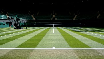 A hyper-realistic, professional photograph from a low angle, looking across the pristine, freshly mown striped lawn of a tennis court. The early morning sun casts long, dramatic shadows. In the background, the iconic dark green stands are empty and quiet, creating a sense of reverence and anticipation. The style should be atmospheric and detailed, evoking the prestige of British sport.