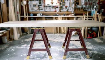 A hyper-realistic photograph in the style of a classic British workshop manual. A pair of sturdy, modern steel sawhorses stands on a clean concrete floor, supporting a beautifully grained piece of oak. Sawdust is lightly scattered around. Soft, natural light streams in from a nearby window, highlighting the tools on a pegboard in the background. The mood is one of craftsmanship, potential, and readiness.