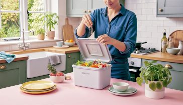 A hyper-realistic, professional photograph in the style of a Gardener's World magazine feature. The image captures a cosy, modern British kitchen with muted green cabinets and a wooden worktop. In the centre, a person's hands are sprinkling light-brown Bokashi bran over fresh food scraps in a clean, stylish grey Bokashi bin. The bin is open, showing layers of colourful vegetable peels. Soft, natural light streams in from a nearby window overlooking a lush, green garden. The mood is clean, eco-conscious, and inspiring.